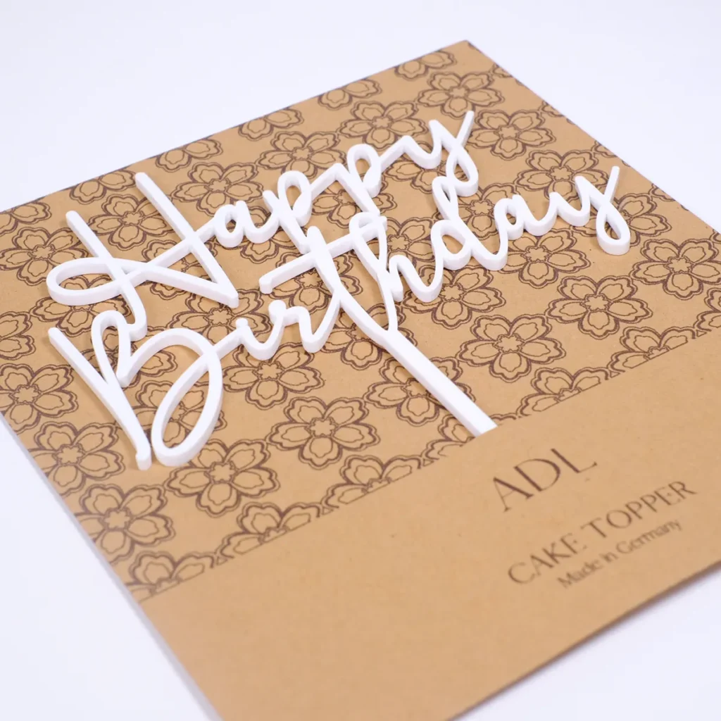 Ein Cake Topper aus weißem Acryl mit Schriftzug "Happy Birthday" in einer braunen Verpackung.