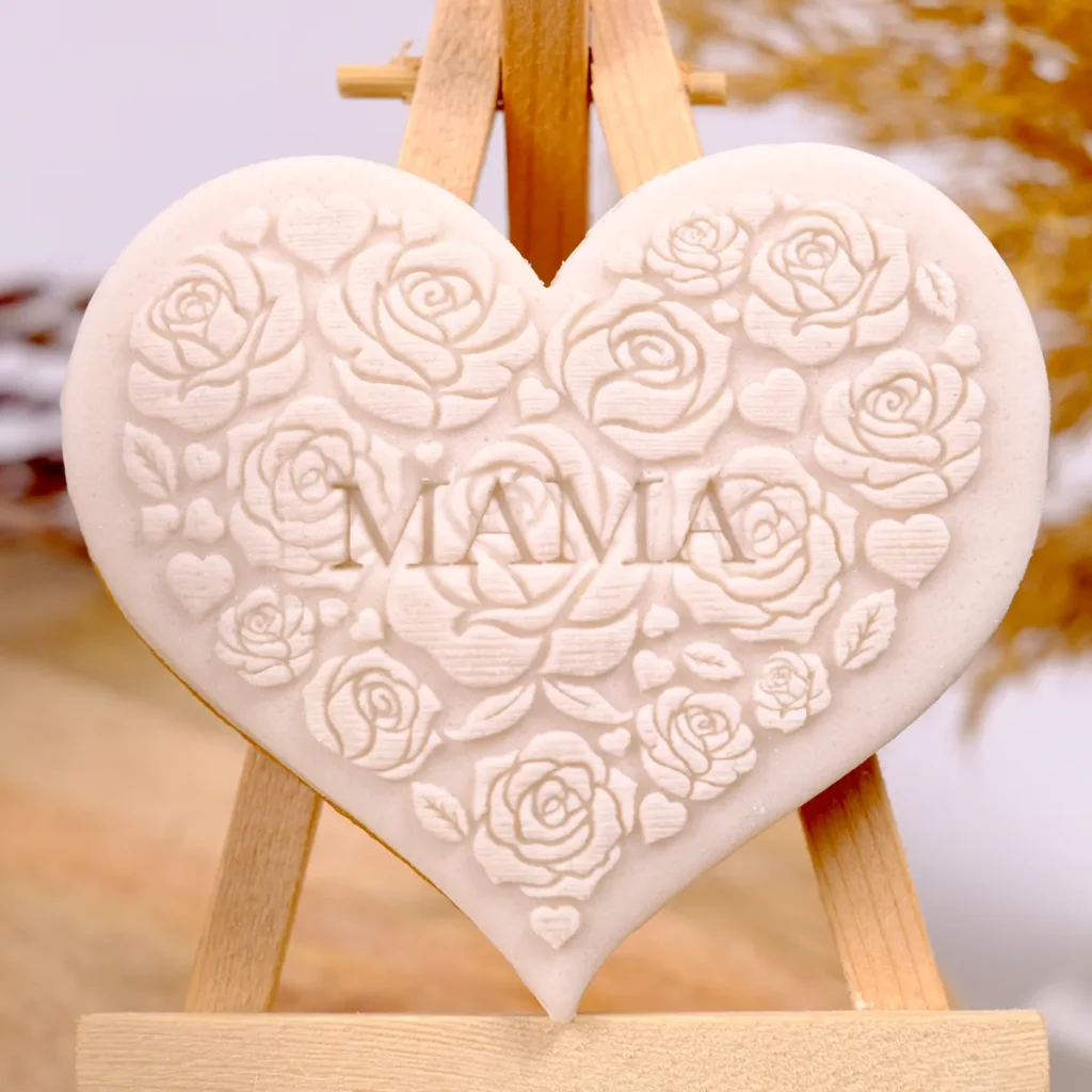Ein beiger Keks in Herzform mit geprägten Rosen und Schriftzug "MAMA" auf einer kleinen Staffelei aus Holz.