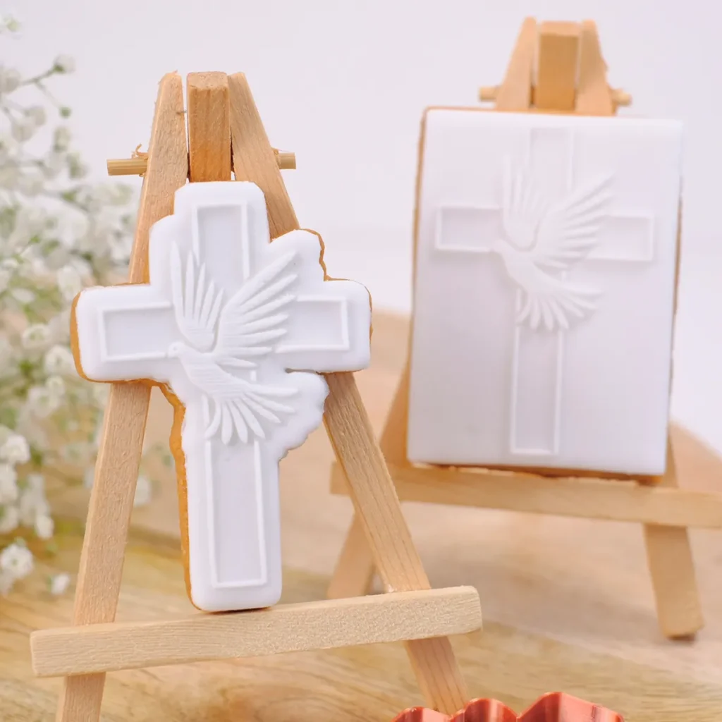 Zwei weiße Kekse mit einem Kreuz und einer Taube auf kleinen Staffeleien aus Holz.