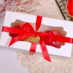 Eine weiße Verpackung mit transparentem Sichtfenster, roter Schleife und zwei Cupcakes auf einer Unterlage aus gehäkeltem Stoff.